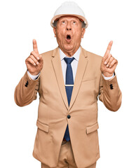 Senior caucasian man wearing architect hardhat amazed and surprised looking up and pointing with fingers and raised arms.