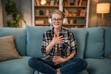 A mature woman listens to a guided meditation on headphones, she is on the couch in her apartment and relaxes - obrazy, fototapety, plakaty