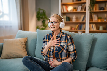 A mature woman listens to a guided meditation on headphones, she is on the couch in her apartment...