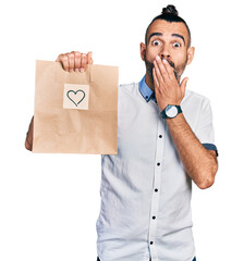 Hispanic man with ponytail holding take away paper bag with heart reminder covering mouth with...