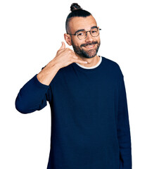 Hispanic man with ponytail wearing casual sweater and glasses smiling doing phone gesture with hand...