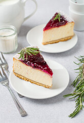Two slices of cheesecake with berry jam, sauce on a white plate, cups, a milk jug, a candle and a sprig of rosemary. Berry cheesecake.