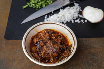 A bowl of slow cooked and braised birria meat in a vintage bowl on a dark brown background with a knife, chopped onions and cilantro