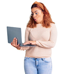 Young latin woman holding laptop scared and amazed with open mouth for surprise, disbelief face