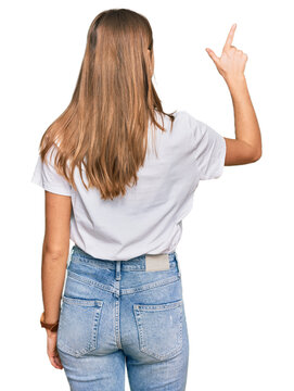 Beautiful young blonde woman wearing casual white t shirt posing backwards pointing ahead with finger hand