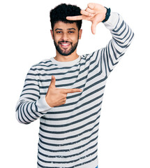 Young arab man with beard wearing casual striped sweater smiling making frame with hands and fingers with happy face. creativity and photography concept.