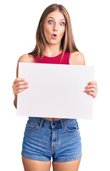 Young beautiful blonde woman holding blank empty banner scared and amazed with open mouth for surprise, disbelief face