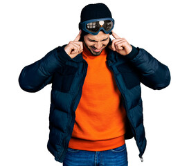 Young hispanic man with beard wearing snow wear and sky glasses covering ears with fingers with annoyed expression for the noise of loud music. deaf concept.