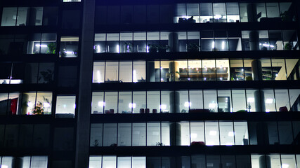 Fragment of the glass facade of a modern corporate building at night. Modern glass office  in city. Big glowing windows in modern office buildings at night, in rows of windows light shines.  - obrazy, fototapety, plakaty