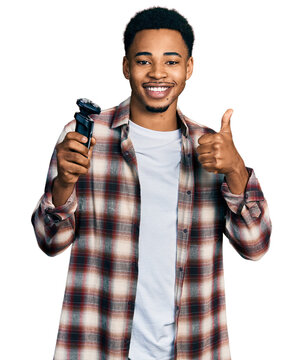 Young african american man holding electric razor machine smiling happy and positive, thumb up doing excellent and approval sign