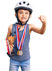 Little cute boy kid wearing bike helmet and winner medals holding winner trophy annoyed and frustrated shouting with anger, yelling crazy with anger and hand raised