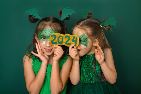 Funny playful girls in green dragon costumes hide behind the golden numbers of the 2024 new year