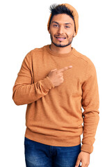Handsome latin american young man wearing winter sweater and wool hat cheerful with a smile of face pointing with hand and finger up to the side with happy and natural expression on face