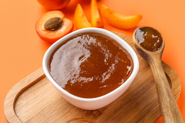 Wooden board with bowl and spoon of sweet apricot jam on orange background