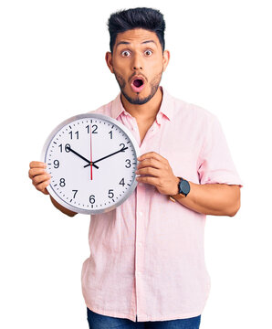 Handsome latin american young man holding big clock scared and amazed with open mouth for surprise, disbelief face