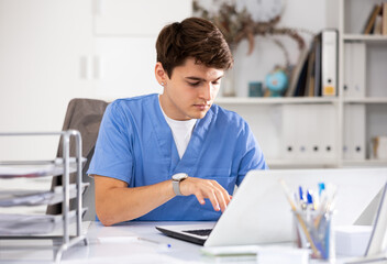 Positive young man doctor therapist sitting in office at workplace and working at laptop