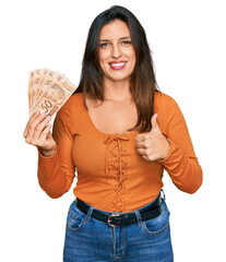 Beautiful hispanic woman holding 50 brazilian real banknotes smiling happy and positive, thumb up...