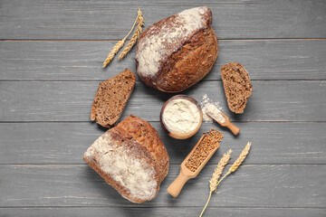 Loaves of fresh rye bread with wheat spikelets, flour and grains on grey wooden background