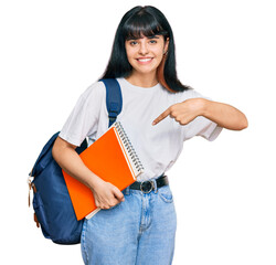 Young hispanic girl wearing student backpack and holding book smiling happy pointing with hand and finger