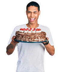 Young hispanic man celebrating birthday holding big chocolate cake winking looking at the camera...