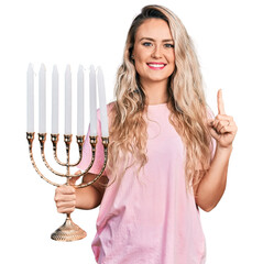Young blonde woman holding menorah hanukkah jewish candle smiling with an idea or question pointing...