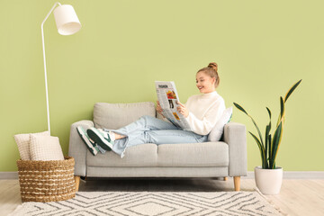 Young woman reading newspaper on sofa at home - obrazy, fototapety, plakaty
