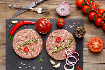 Raw beef hamburger patties on stone plate, vegetables and spices, top view, wooden background.