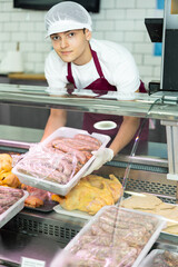 Young salesman puts tray with semi-finished raw sausages for barbecue in refrigerator window. Made-up meat product for frying on grill and open fire - kupaty, sausages