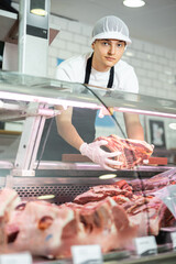 Positive young salesman standing behind counter demonstrating piece of meat in butcher shop