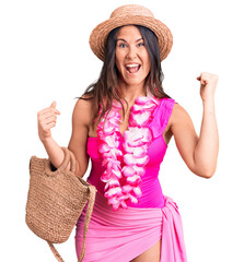 Young beautiful brunette woman wearing swimwear and hawaiian lei holding bag screaming proud,...