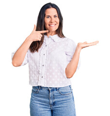 Young beautiful brunette woman wearing casual shirt amazed and smiling to the camera while presenting with hand and pointing with finger.