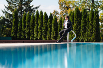 Handsome businessman in suit and with sunglasses sitting on the edge of a swimming pool and looking away at suny summer day
