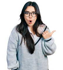 Beautiful hispanic woman wearing casual sweater and glasses surprised pointing with hand finger to...
