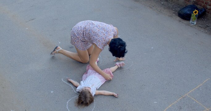 Mother and baby child are playing fun game, drawing with chalk on asphalt. Creating a human figure using live stencil. Playful experience for children and their parents on playground.
