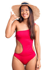 Young beautiful latin girl wearing swimwear and summer hat smiling doing phone gesture with hand and fingers like talking on the telephone. communicating concepts.