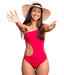 Young beautiful latin girl wearing swimwear and summer hat looking at the camera smiling with open...