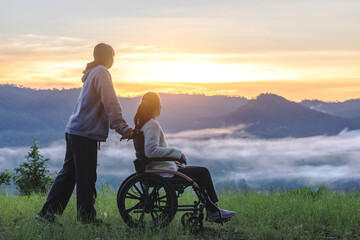 Silhouette of woman in wheelchair with care helper walking around in spring nature at sunrise misty mountains. - obrazy, fototapety, plakaty