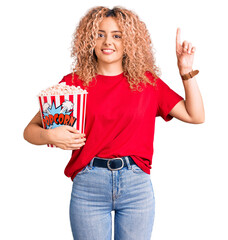 Young blonde woman with curly hair eating popcorn surprised with an idea or question pointing finger with happy face, number one