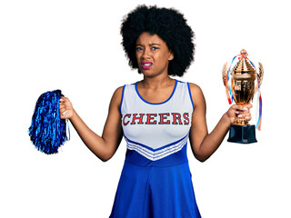 Young african american woman wearing cheerleader uniform holding pompom and trophy clueless and...