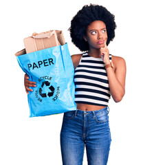Young african american woman holding recycling bag with paper and cardboard serious face thinking...