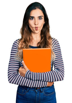 Beautiful hispanic woman holding book making fish face with mouth and squinting eyes, crazy and comical.