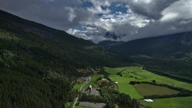 Aerial dolly shot over Innerdalen valley with rural road, farm buildings and meadows surrounded by vast forests, mountain ridge and peak covered in white clouds. Norway nature travel concept