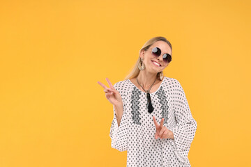 Portrait of smiling hippie woman showing peace signs on yellow background. Space for text