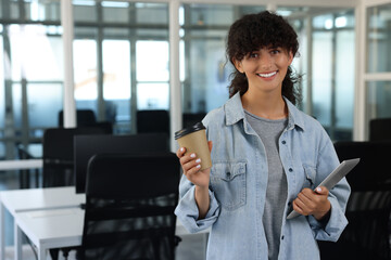 Happy woman with paper cup of drink and tablet in office, space for text