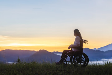 Woman is sitting on wheelchair looking sunset mountains background.