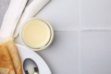 Tasty condensed milk in jar and crepes on white tiled table, flat lay. Space for text