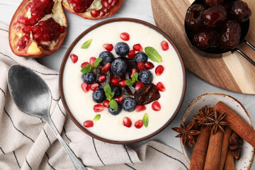 Delicious semolina pudding with blueberries, pomegranate, dates and mint in bowl served on white table, flat lay