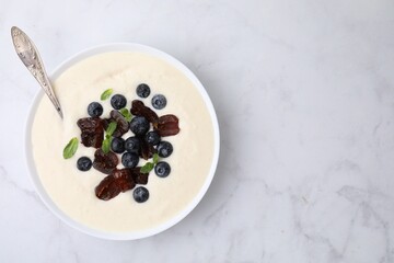 Delicious semolina pudding with blueberries, dates, mint and spoon in bowl on white marble table, top view. Space for text