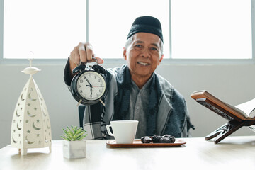 Elderly Asian muslim man holding alarm clock showing iftar time while sitting on dining table