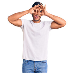 Young latin man wearing casual clothes doing ok gesture like binoculars sticking tongue out, eyes looking through fingers. crazy expression.